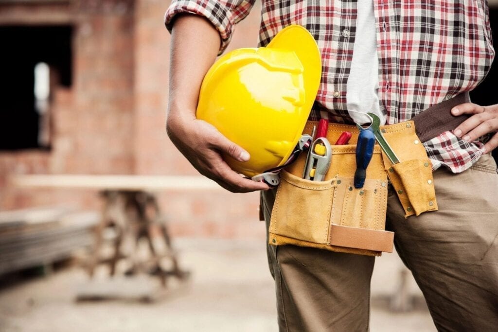 A worker holding his yellow construction cap and tools tied to his waist